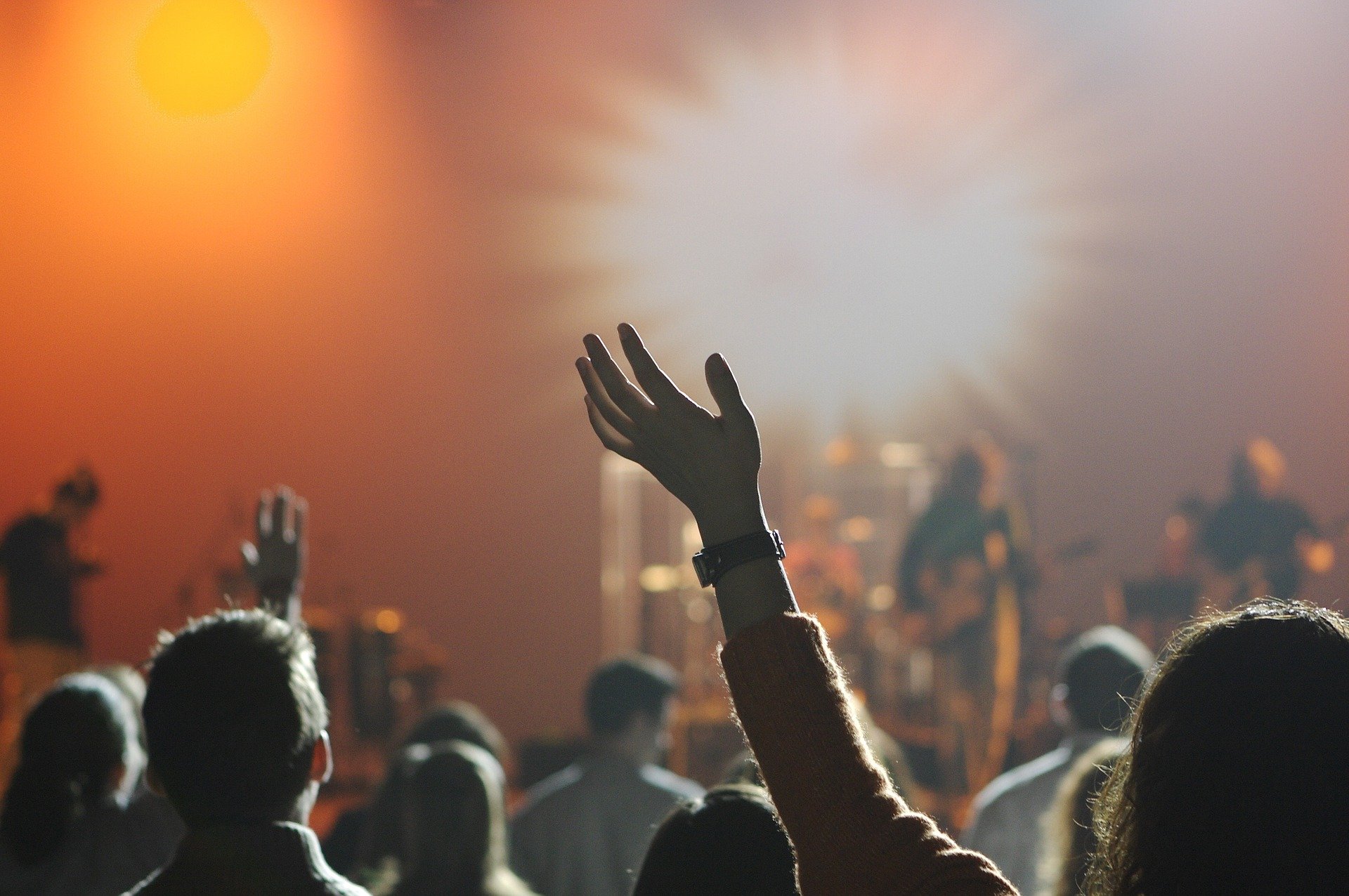 Image of people in the crowd at a concert, stage is out of focus, women's hand raised and waving to the music is main focus of the frame.