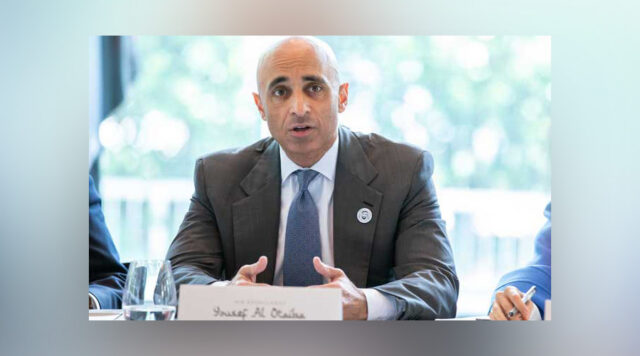 Ambassador Yousef Al Otaiba in a gray suit, white dress shirt and blue tie at a table addressing the audience